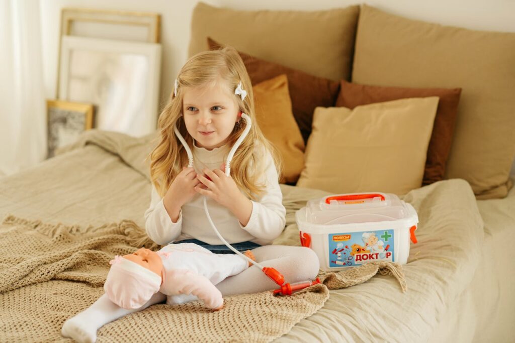 A cute child with blonde hair playing doctor on a bed with a toy stethoscope and baby doll.